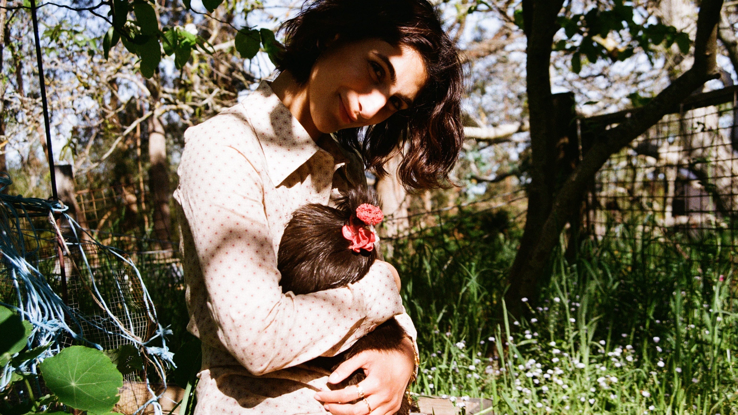 Woman holding a chicken in a natural outdoor setting with trees and greenery.